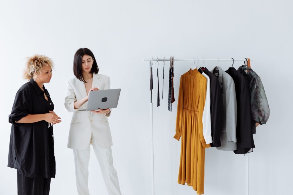 Two women reviewing the Latest seasonal style trends on a laptop near a clothes rack.